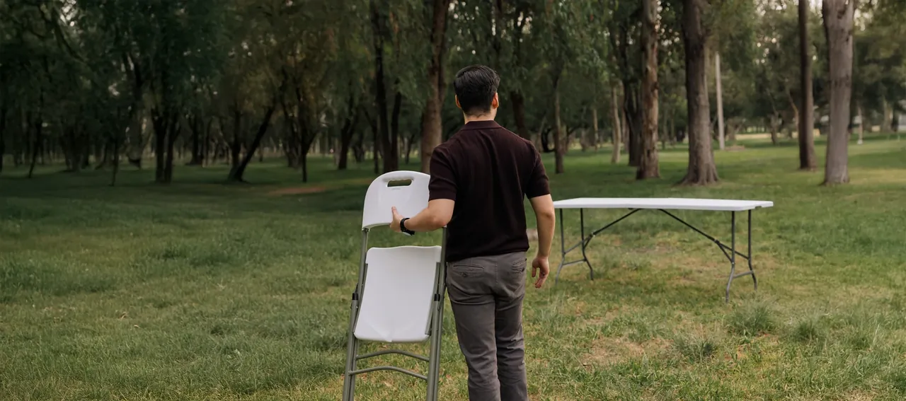 Persona llevando una silla plegable hacia una mesa en un jardín exterior